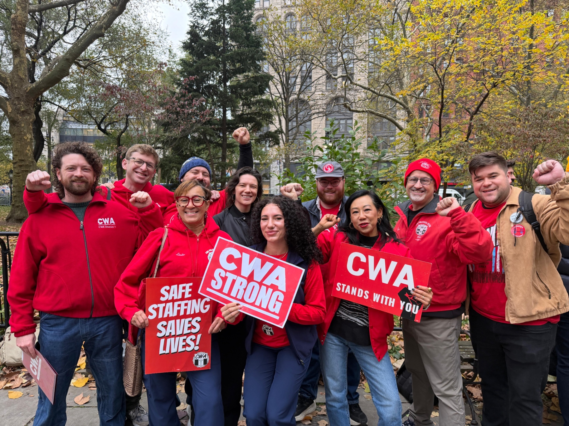 Nurses in red CWA gear holding Safe Staffing Saves Lives and CWA Strong signs in a park.